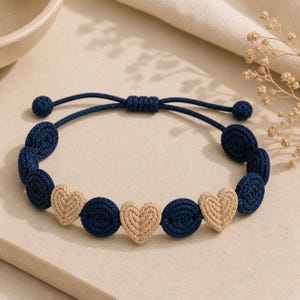 Peut inclure: Un bracelet en crochet bleu marine et beige. Le bracelet présente des perles circulaires bleu marine et des perles en forme de cœur beiges alternées. Le cordon réglable est bleu marine.
