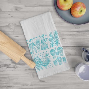 May include: A white kitchen towel featuring a teal blue folk-art print of a rooster, stalks of corn, wheat sheaves, and two figures standing near a basket. The towel is positioned next to a wooden rolling pin, a grey plate with two apples, and two mugs.