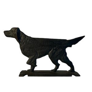 May include: A dark brown, cast metal figurine of a standing dog in profile. The dog is depicted with its head turned slightly to the left, a long tail extended horizontally, and textured fur details throughout its body, mounted on a flat base.
