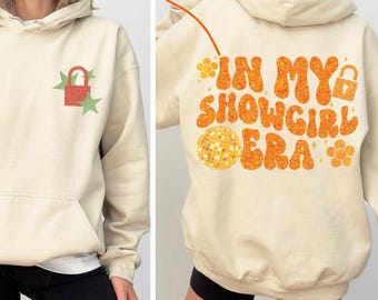 May include: Cream-colored hoodie with a front pocket and hood. The front features a red lock and two green stars. The back displays the text "IN MY SHOWGIRL ERA" in orange, along with a disco ball and other decorative elements. A casual, comfortable garment.