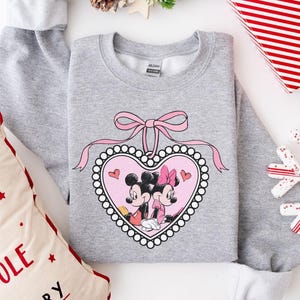 Op de afbeelding: Een grijze sweatshirt met een roze hartontwerp met Mickey en Minnie Mouse. Het hart is omrand met witte parels en een roze strik. De sweatshirt is omgeven door feestelijke decoraties, waaronder een kussen en een sneeuwvlok.