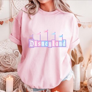 May include: A pink t-shirt with a pastel-colored graphic of the Disneyland castle and the word "Disneyland" in a blocky font.