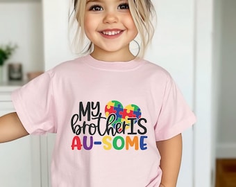May include: A young girl wearing a pink t-shirt with the text "My brother is AU-SOME" and a colorful puzzle piece heart graphic.