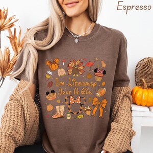 Puede incluir: Camiseta color espresso con el texto "I'm Literally Just A Girl" y gráficos con temática otoñal. El diseño incluye calabazas, un balón de fútbol americano, un lazo y una magdalena de Minnie Mouse. La camiseta se combina con una rebeca marrón y pantalones negros.