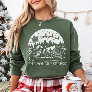 Puede incluir: Una sudadera verde con un gráfico blanco de un tren y las palabras "The Polar Express". La persona que lleva la sudadera está sentada en una manta de cuadros rojos y blancos y sostiene una taza blanca con una ilustración de Papá Noel.