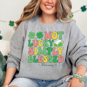 Puede incluir: Una sudadera gris con un gr&aacute;fico verde, amarillo y rosa que dice "Not Lucky Simply Blessed Romans 4:7-8".