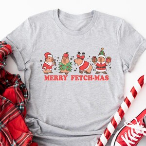 May include: A gray t-shirt with a Christmas-themed design featuring four capybaras dressed in Santa hats and holiday attire. The text "Merry Fetch-Mas" is printed below the capybaras.