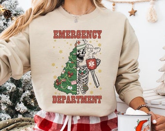 Puede incluir: Una sudadera beige con un gráfico navideño de un esqueleto con un gorro de Papá Noel y sosteniendo una taza. El gráfico dice "Emergency Department". La sudadera la lleva puesta una persona con pantalones de cuadros rojos y blancos.