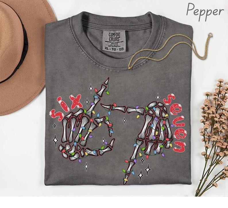 May include: A dark grey t-shirt with a skeleton hand design. The hands are decorated with colorful Christmas lights and the words "Six" and "Seven" in red. A gold necklace is draped across the shirt.