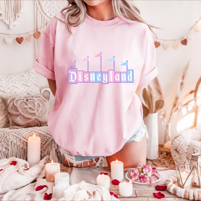 May include: A pink t-shirt with a pastel-colored graphic of the Disneyland castle and the word "Disneyland" in a blocky font.