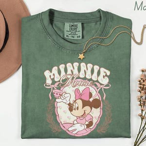 Puede incluir: Una camiseta verde musgo con un dise&ntilde;o de Minnie Mouse. El dise&ntilde;o muestra a Minnie Mouse con una taza de t&eacute;, rodeada de flores y las palabras "Minnie Mouse". Un collar dorado con un colgante de estrella descansa sobre la camiseta.