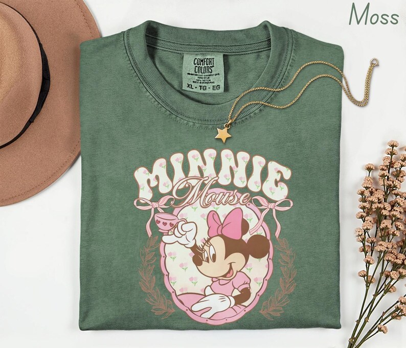Puede incluir: Una camiseta verde musgo con un dise&ntilde;o de Minnie Mouse. El dise&ntilde;o muestra a Minnie Mouse con una taza de t&eacute;, rodeada de flores y las palabras "Minnie Mouse". Un collar dorado con un colgante de estrella descansa sobre la camiseta.