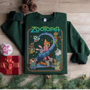 Pu&ograve; includere: Una felpa verde scuro con la scritta "Zootopia" e una grafica colorata dei personaggi del film d'animazione. La felpa &egrave; circondata da decorazioni natalizie, tra cui una scatola regalo rossa, una palla bianca e rami di pino.