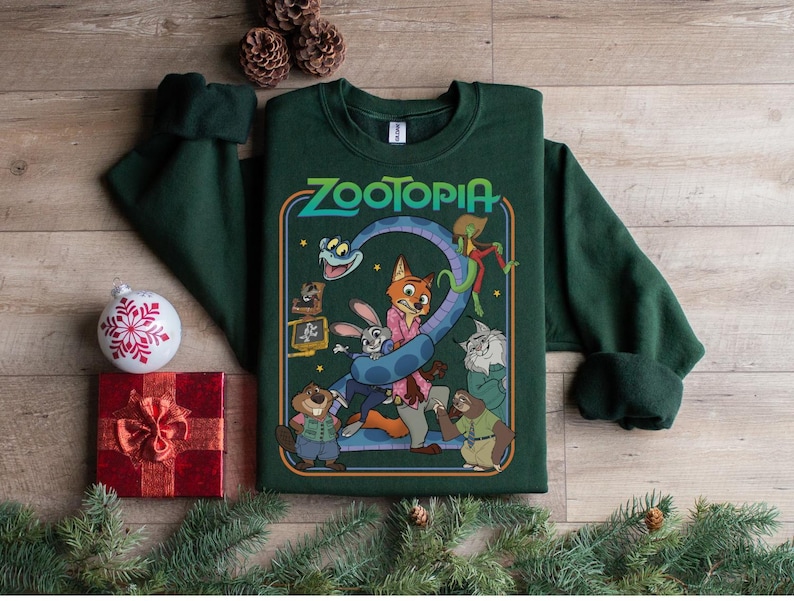 Pu&ograve; includere: Una felpa verde scuro con la scritta "Zootopia" e una grafica colorata dei personaggi del film d'animazione. La felpa &egrave; circondata da decorazioni natalizie, tra cui una scatola regalo rossa, una palla bianca e rami di pino.