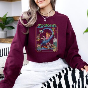 Pu&ograve; includere: Una felpa bordeaux con una grafica colorata del film d'animazione "Zootropolis". La grafica include il titolo del film e vari personaggi. La felpa &egrave; a maniche lunghe e girocollo.