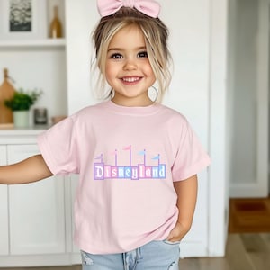 May include: A young girl wearing a pink t-shirt with a pastel blue and pink Disneyland graphic. She is wearing a light blue bow in her hair.