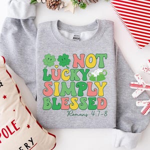 Puede incluir: Sudadera gris con un dise&ntilde;o de tr&eacute;bol verde y el texto "Not Lucky Simply Blessed Romans 4:7-8".