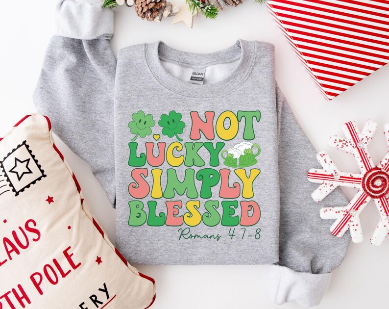 Puede incluir: Sudadera gris con un dise&ntilde;o de tr&eacute;bol verde y el texto "Not Lucky Simply Blessed Romans 4:7-8".