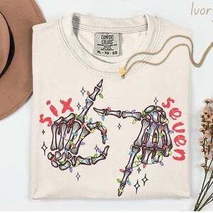 May include: Ivory-colored t-shirt with a skeleton hand graphic. The graphic features two skeleton hands forming the number six and seven with colorful Christmas lights. The words "six" and "seven" are in red.
