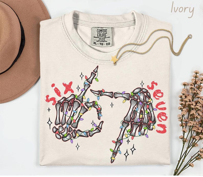 May include: Ivory-colored t-shirt with a skeleton hand graphic. The graphic features two skeleton hands forming the number six and seven with colorful Christmas lights. The words "six" and "seven" are in red.