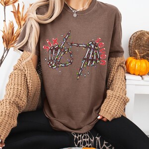May include: Espresso-colored t-shirt featuring a skeleton hand graphic with the text "Six Senses" in red. The skeleton hands are decorated with colorful Christmas lights. The shirt is a casual, comfortable style.