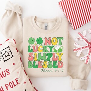 Puede incluir: Una sudadera beige con un gr&aacute;fico verde, amarillo, rosa y naranja que dice "Not Lucky Simply Blessed" con un tr&eacute;bol y una jarra de cerveza. El texto "Romanos 4:7-8" est&aacute; debajo del gr&aacute;fico.