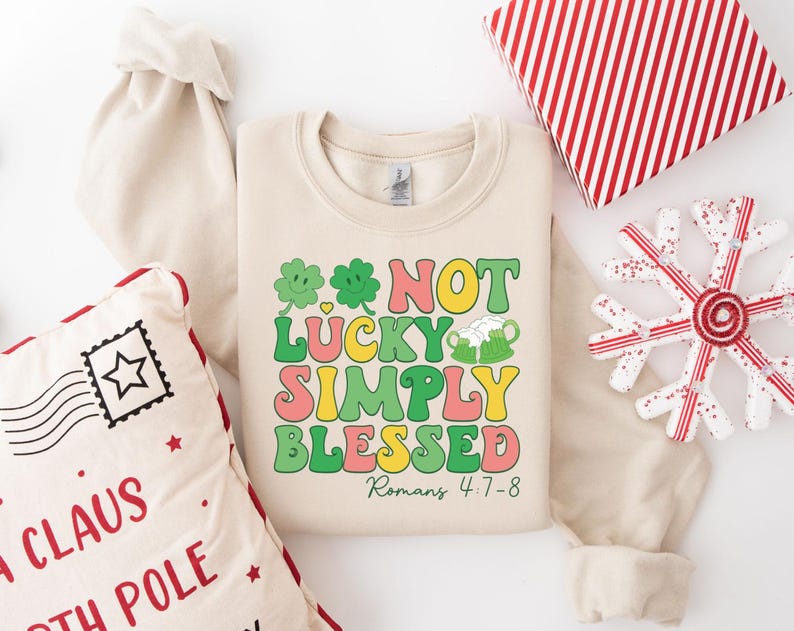 Puede incluir: Una sudadera beige con un gr&aacute;fico verde, amarillo, rosa y naranja que dice "Not Lucky Simply Blessed" con un tr&eacute;bol y una jarra de cerveza. El texto "Romanos 4:7-8" est&aacute; debajo del gr&aacute;fico.