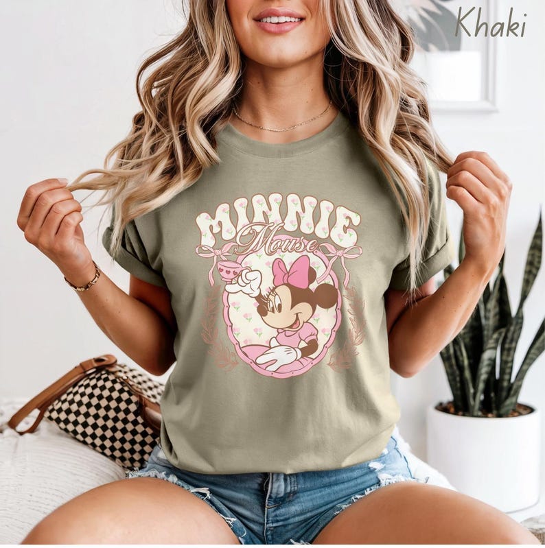Puede incluir: Camiseta caqui con un gr&aacute;fico de Minnie Mouse. El dise&ntilde;o presenta a Minnie Mouse con un vestido y lazo rosa, sosteniendo una taza de t&eacute;. Las palabras "Minnie Mouse" est&aacute;n arqueadas sobre la imagen en una fuente retro.