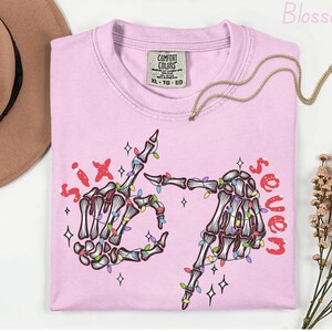May include: A light pink t-shirt with a skeleton hand graphic. The graphic includes the words "six" and "seven" in red, and the skeleton hands are decorated with colorful string lights. A gold necklace is draped across the shirt.