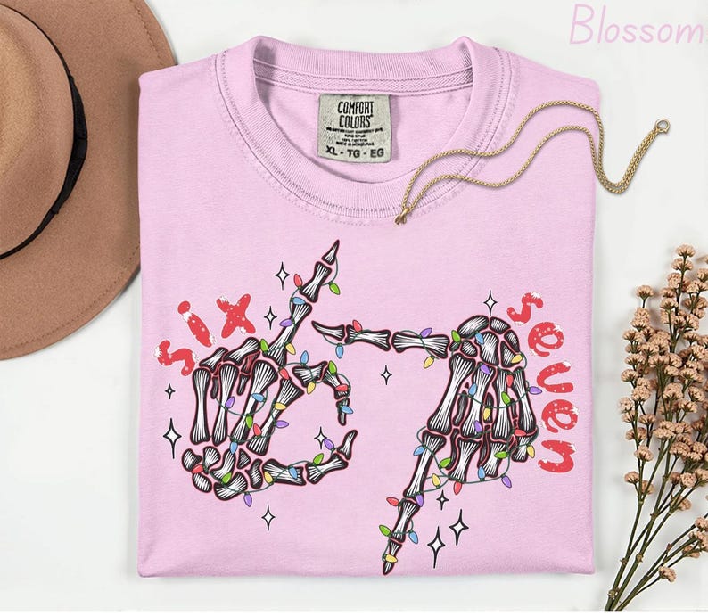 May include: A light pink t-shirt with a skeleton hand graphic. The graphic includes the words "six" and "seven" in red, and the skeleton hands are decorated with colorful string lights. A gold necklace is draped across the shirt.