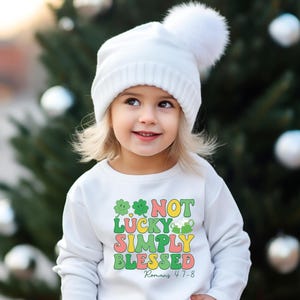 Puede incluir: Una sudadera blanca con un tr&eacute;bol verde y el texto "Not Lucky Simply Blessed Romans 4:7-8" en letras de colores.