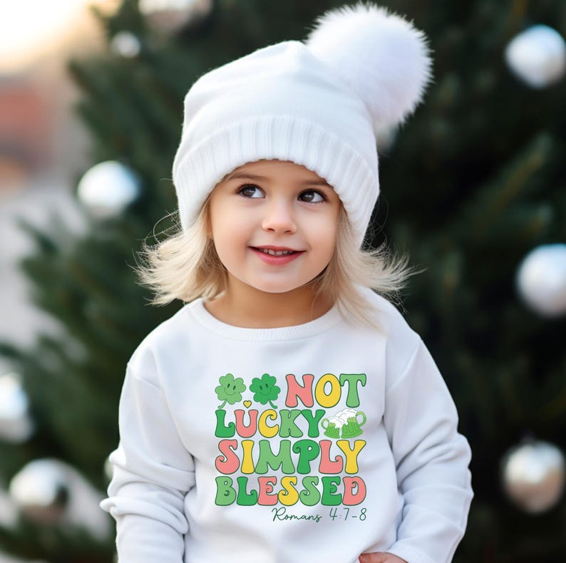 Puede incluir: Una sudadera blanca con un tr&eacute;bol verde y el texto "Not Lucky Simply Blessed Romans 4:7-8" en letras de colores.