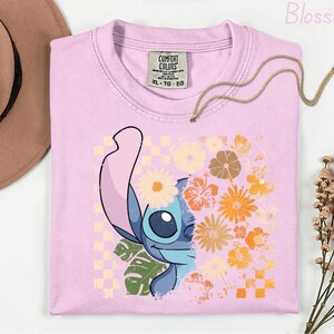 Puede incluir: Camiseta rosa claro Comfort Colors con un gr&aacute;fico de Stitch y detalles florales. El dise&ntilde;o incorpora un patr&oacute;n de tablero de ajedrez y flores naranjas, amarillas y blancas. Tambi&eacute;n se ve un collar dorado y un sombrero marr&oacute;n.