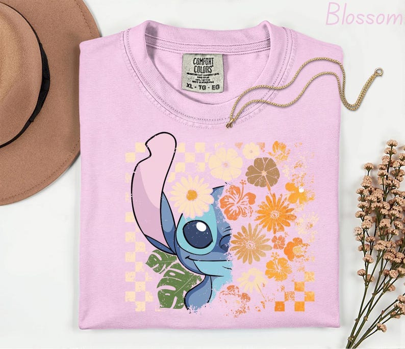 Puede incluir: Camiseta rosa claro Comfort Colors con un gr&aacute;fico de Stitch y detalles florales. El dise&ntilde;o incorpora un patr&oacute;n de tablero de ajedrez y flores naranjas, amarillas y blancas. Tambi&eacute;n se ve un collar dorado y un sombrero marr&oacute;n.