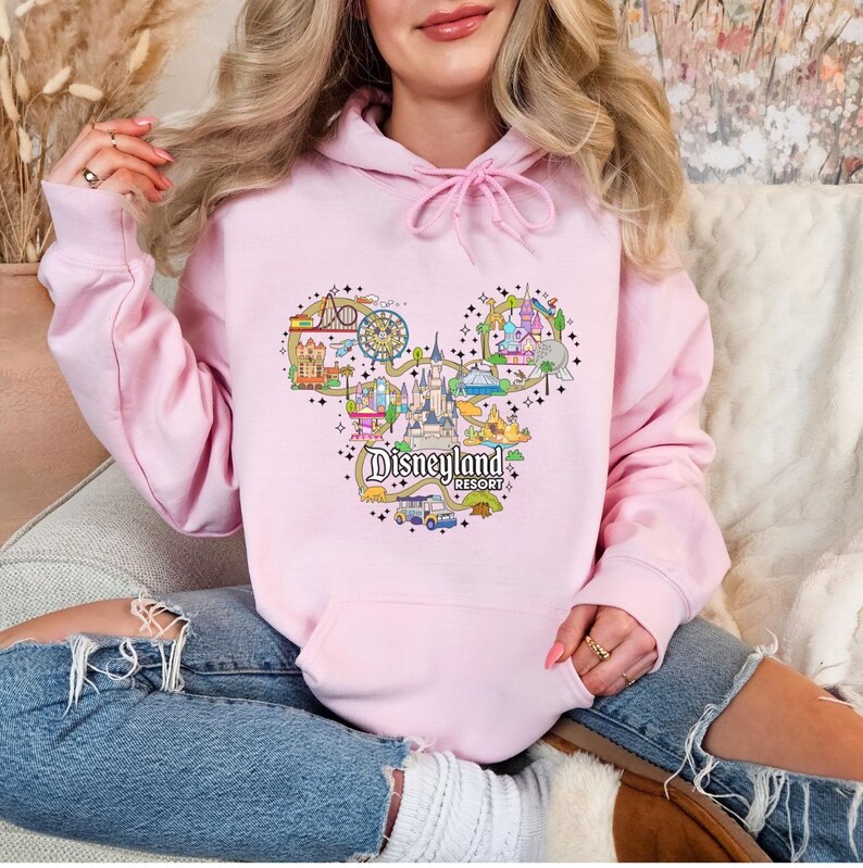 May include: A pale pink hoodie featuring a Disneyland Resort design in the shape of Mickey Mouse's head. The graphic includes colourful illustrations of park attractions and the Disneyland Resort logo.