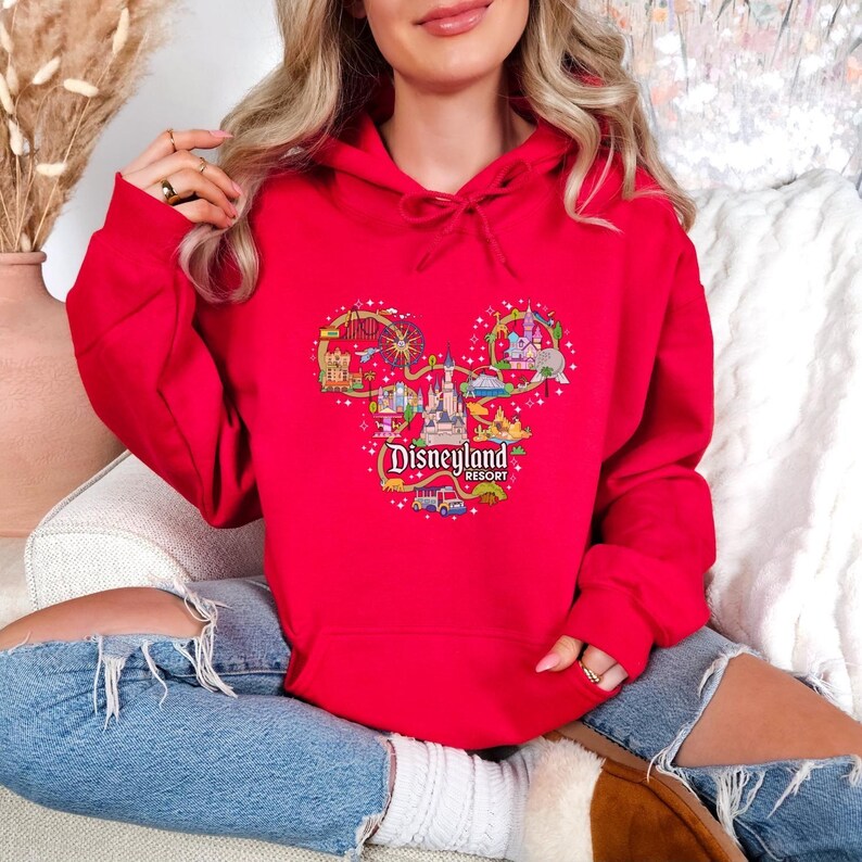 May include: A red hoodie with a Disneyland Resort graphic in the shape of Mickey Mouse's head. The graphic features illustrations of park attractions and the Disneyland Resort logo. The hoodie is made of a soft material.