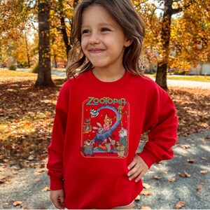 Pu&ograve; includere: Una felpa rossa con la scritta "Zootopia" in verde e una grafica colorata dei personaggi del film d'animazione. La felpa &egrave; a maniche lunghe ed &egrave; indossata da una persona giovane.