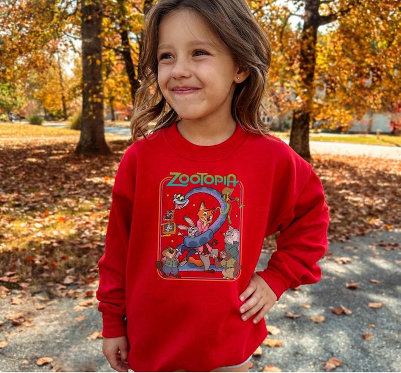 Pu&ograve; includere: Una felpa rossa con la scritta "Zootopia" in verde e una grafica colorata dei personaggi del film d'animazione. La felpa &egrave; a maniche lunghe ed &egrave; indossata da una persona giovane.