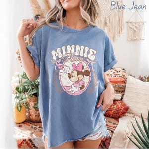 Puede incluir: Camiseta azul vaquera con un gr&aacute;fico de Minnie Mouse. El gr&aacute;fico incluye el texto "MINNIE Mouse" en una fuente vintage, junto con una ilustraci&oacute;n de Minnie Mouse con un vestido y lazo rosa. La camiseta tiene mangas enrolladas.