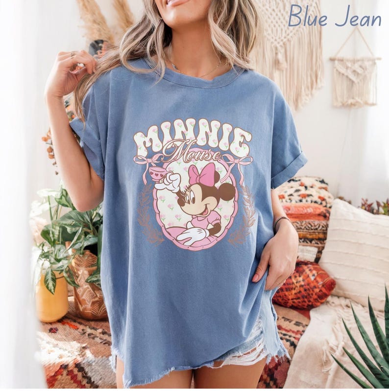 Puede incluir: Camiseta azul vaquera con un gr&aacute;fico de Minnie Mouse. El gr&aacute;fico incluye el texto "MINNIE Mouse" en una fuente vintage, junto con una ilustraci&oacute;n de Minnie Mouse con un vestido y lazo rosa. La camiseta tiene mangas enrolladas.