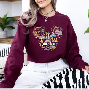 May include: Burgundy sweatshirt with a Mickey Mouse head design filled with Disneyland Resort imagery. The design includes a castle, Ferris wheel, and other park attractions. The text "Disneyland Resort" is also visible.