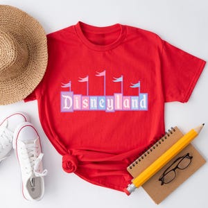 May include: Red t-shirt with a graphic of the Disneyland logo in colorful blocks with flags on top. The logo is in a vintage style.