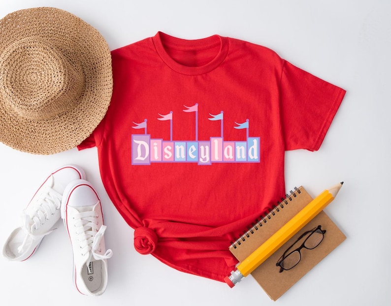 May include: Red t-shirt with a graphic of the Disneyland logo in colorful blocks with flags on top. The logo is in a vintage style.