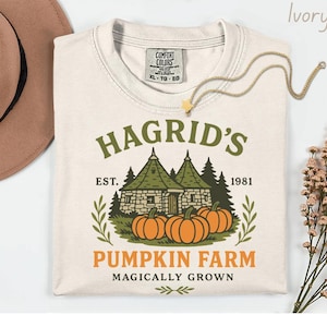 Peut inclure: T-shirt ivoire avec un motif de la Ferme aux Potirons de Hagrid. Le design comprend un bâtiment, des citrouilles et le texte "Hagrid's Pumpkin Farm Magically Grown". L'année 1981 est également présente.