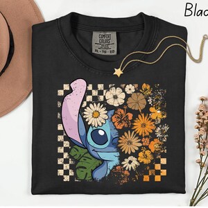 Puede incluir: Camiseta negra con un dise&ntilde;o de personaje de dibujos animados y floral. El dise&ntilde;o presenta margaritas, hibiscos y otras flores en tonos naranja, amarillo y blanco. Un collar dorado con un colgante de estrella est&aacute; drapeado sobre la camiseta.