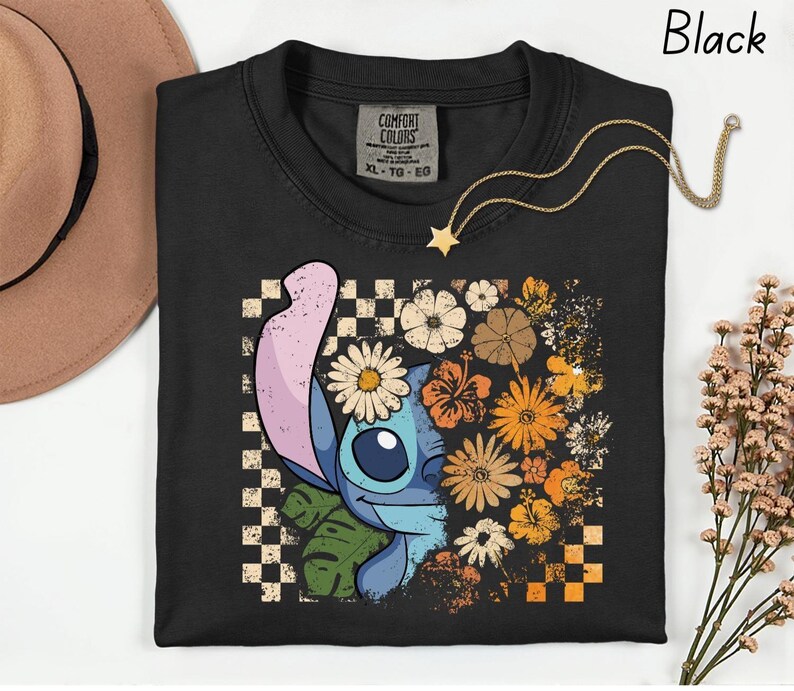 Puede incluir: Camiseta negra con un dise&ntilde;o de personaje de dibujos animados y floral. El dise&ntilde;o presenta margaritas, hibiscos y otras flores en tonos naranja, amarillo y blanco. Un collar dorado con un colgante de estrella est&aacute; drapeado sobre la camiseta.