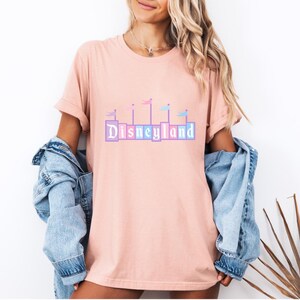 May include: A pink t-shirt with a graphic of the Disneyland castle with flags in pastel colors. The text "DISNEYLAND" is printed in colorful blocks.