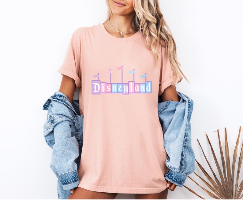 May include: A pink t-shirt with a graphic of the Disneyland castle with flags in pastel colors. The text "DISNEYLAND" is printed in colorful blocks.