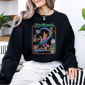 Pu&ograve; includere: Felpa girocollo nera con grafica colorata del film d'animazione "Zootopia". Il design include il titolo del film e vari personaggi, come una volpe, un coniglio e altri animali. La felpa &egrave; uno stile casual e confortevole.