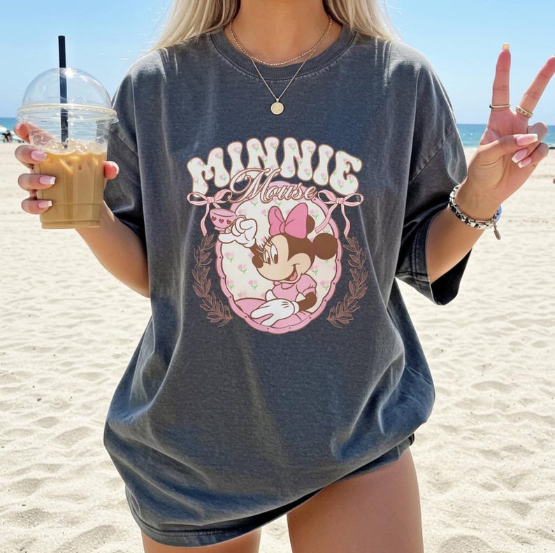 Puede incluir: Camiseta gris oscuro con un gr&aacute;fico de Minnie Mouse. El dise&ntilde;o incluye el texto "Minnie Mouse" en una fuente retro, un marco floral rosa y una ilustraci&oacute;n de Minnie Mouse sosteniendo una taza de t&eacute;. La camiseta es de gran tama&ntilde;o.